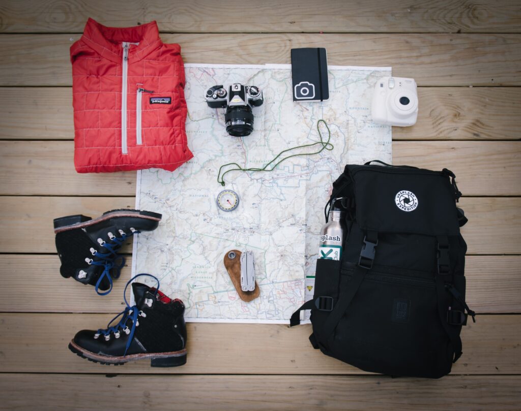 Holiday packing list items, coat, boots, backpack, Multi tool, compass, camera, notepad and map on a wooden floor